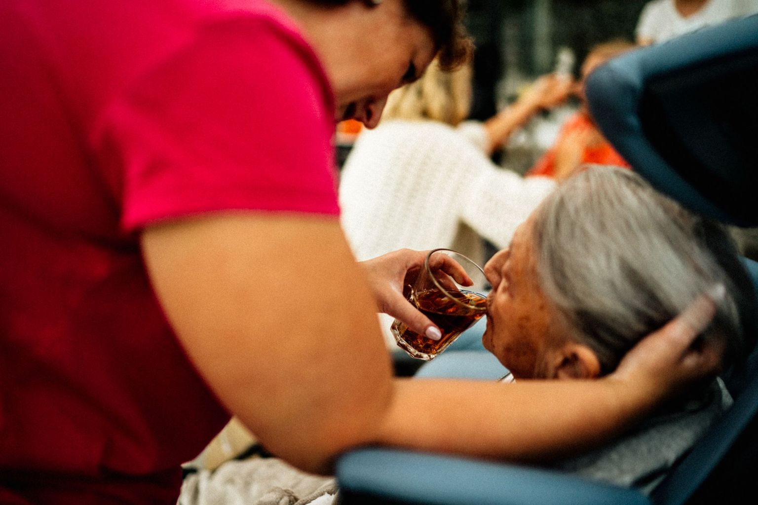A caregiver gently helps an older woman drink tea.