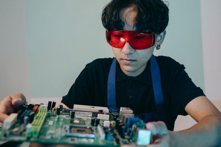 A trainee in safety glasses works on a circuit board.